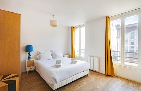 Une chambre à coucher avec un grand lit, une table de chevet avec une lampe, une fenêtre avec des rideaux et un radiateur.