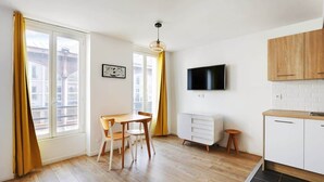 Een moderne keuken met houten kastjes, een eethoek met een tafel en stoelen, en een aan de muur bevestigde televisie.