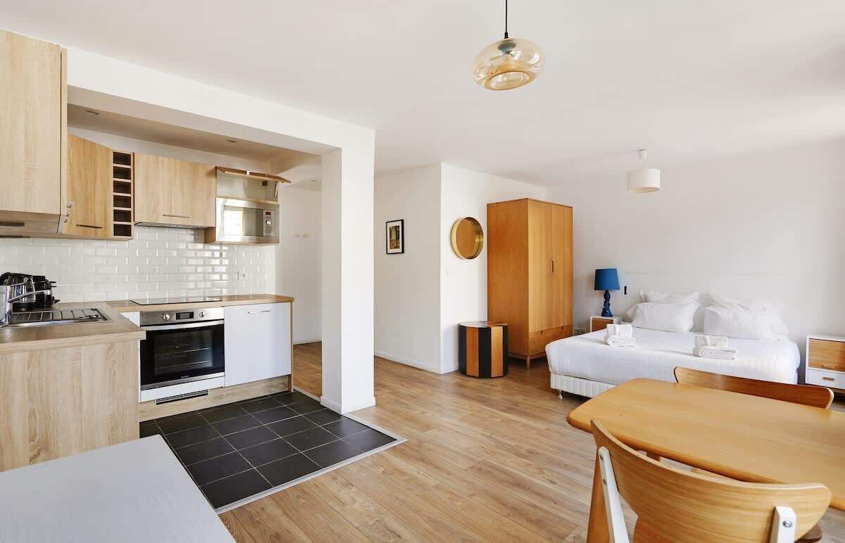 Cozinha moderna com armários de madeira, micro-ondas e fogão. Há uma área de jantar com mesa e cadeiras, e um quarto com cama e mesa de cabeceira.