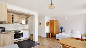 Een moderne keuken met houten kastjes, een magnetron en een kookplaat. Er is een eethoek met een tafel en stoelen, en een slaapkamer met een bed en nachtkastje.