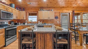A kitchen with wooden cabinets, granite countertops, a central island with bar stools, and a dining area with a wooden table and chairs. - Gorgeous Views, Game Room, Sauna, True Log Cabin! | Free Attractions! (Gatlinburg)