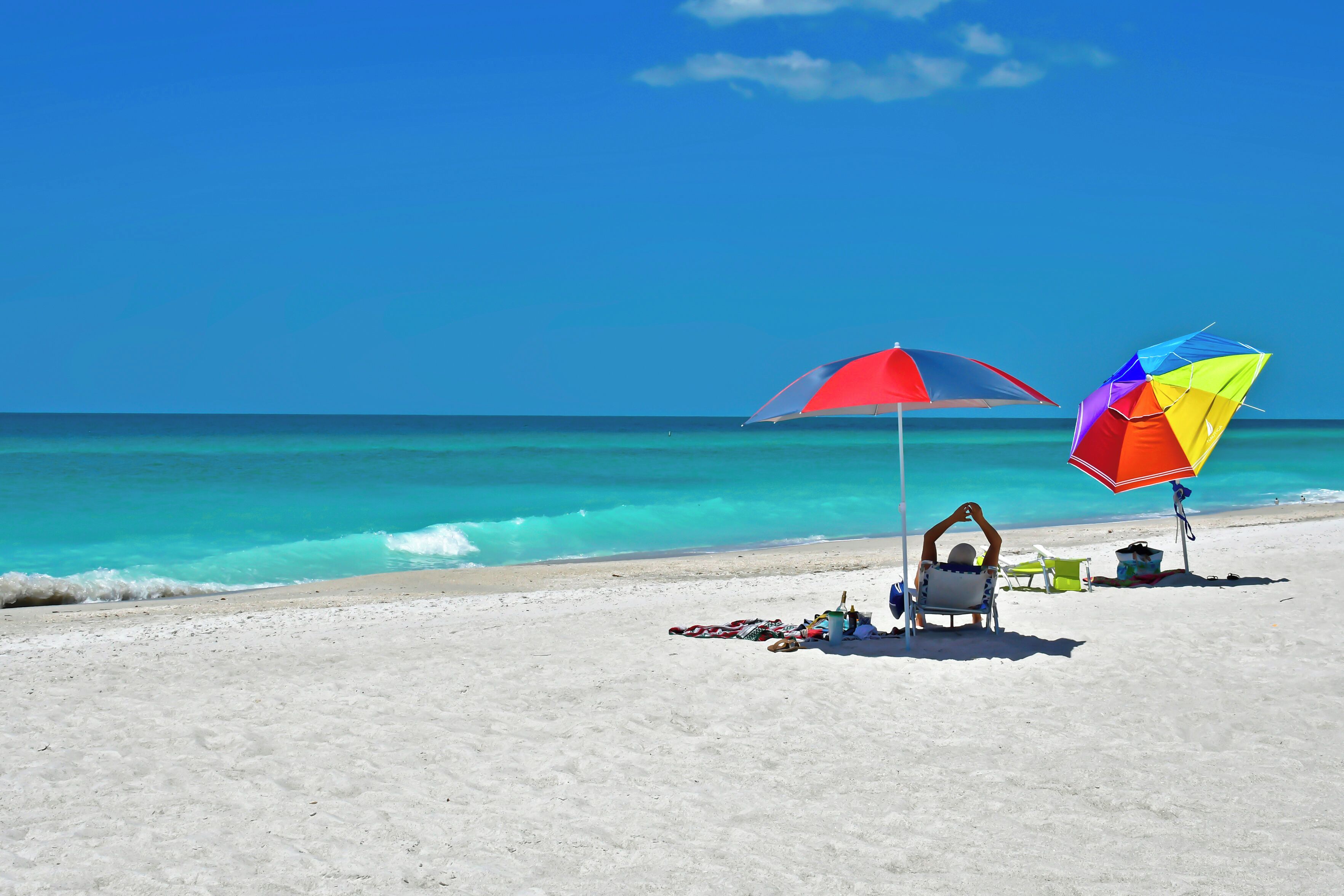 A beach with two colorful umbrellas, a chair, and a small table with items on the sand.