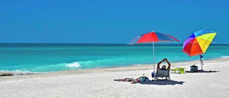 A beach with two colorful umbrellas, a chair, and a small table with items on the sand.