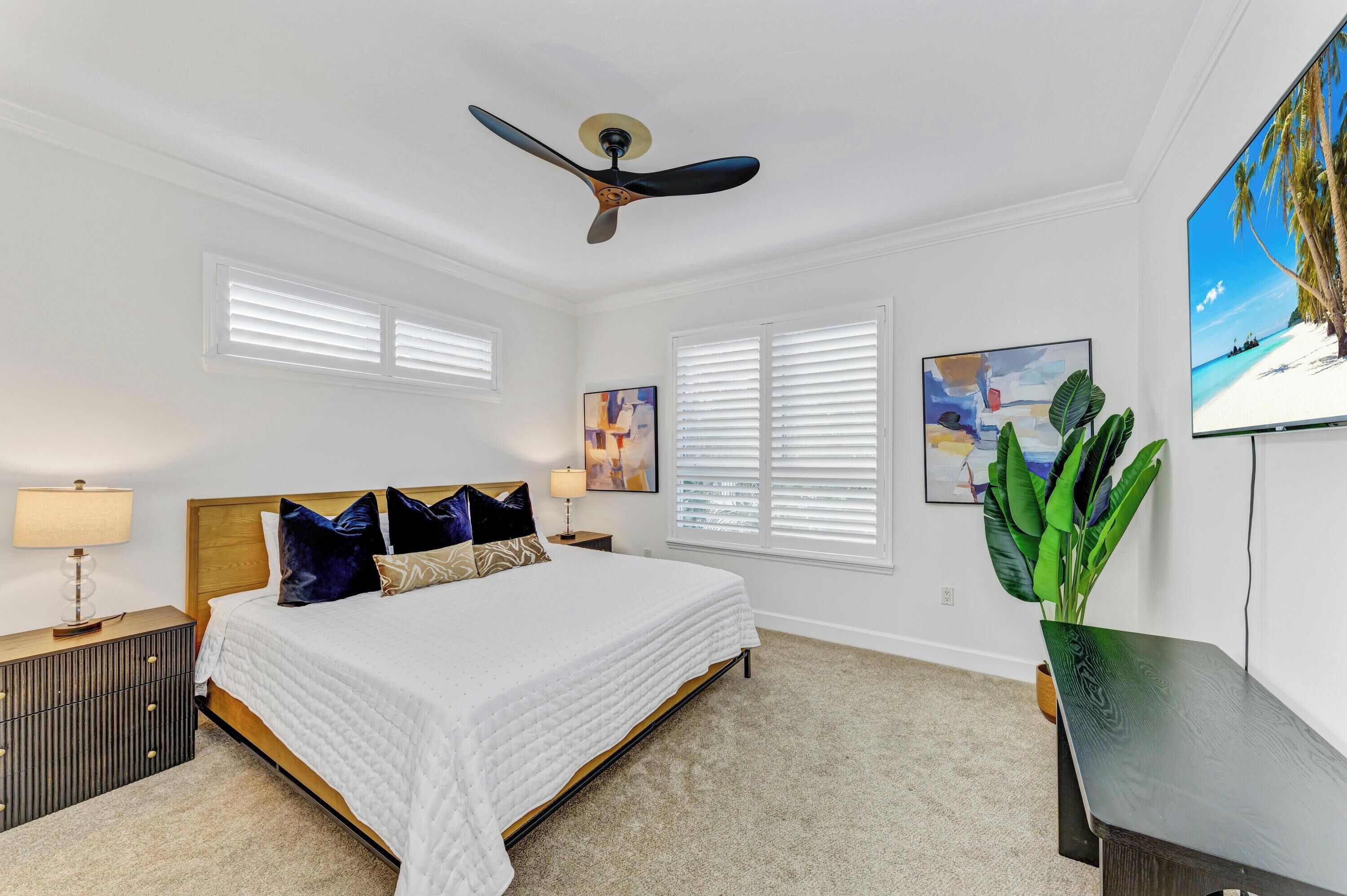 A bedroom with a bed, a ceiling fan, a TV, and a large window with blinds.