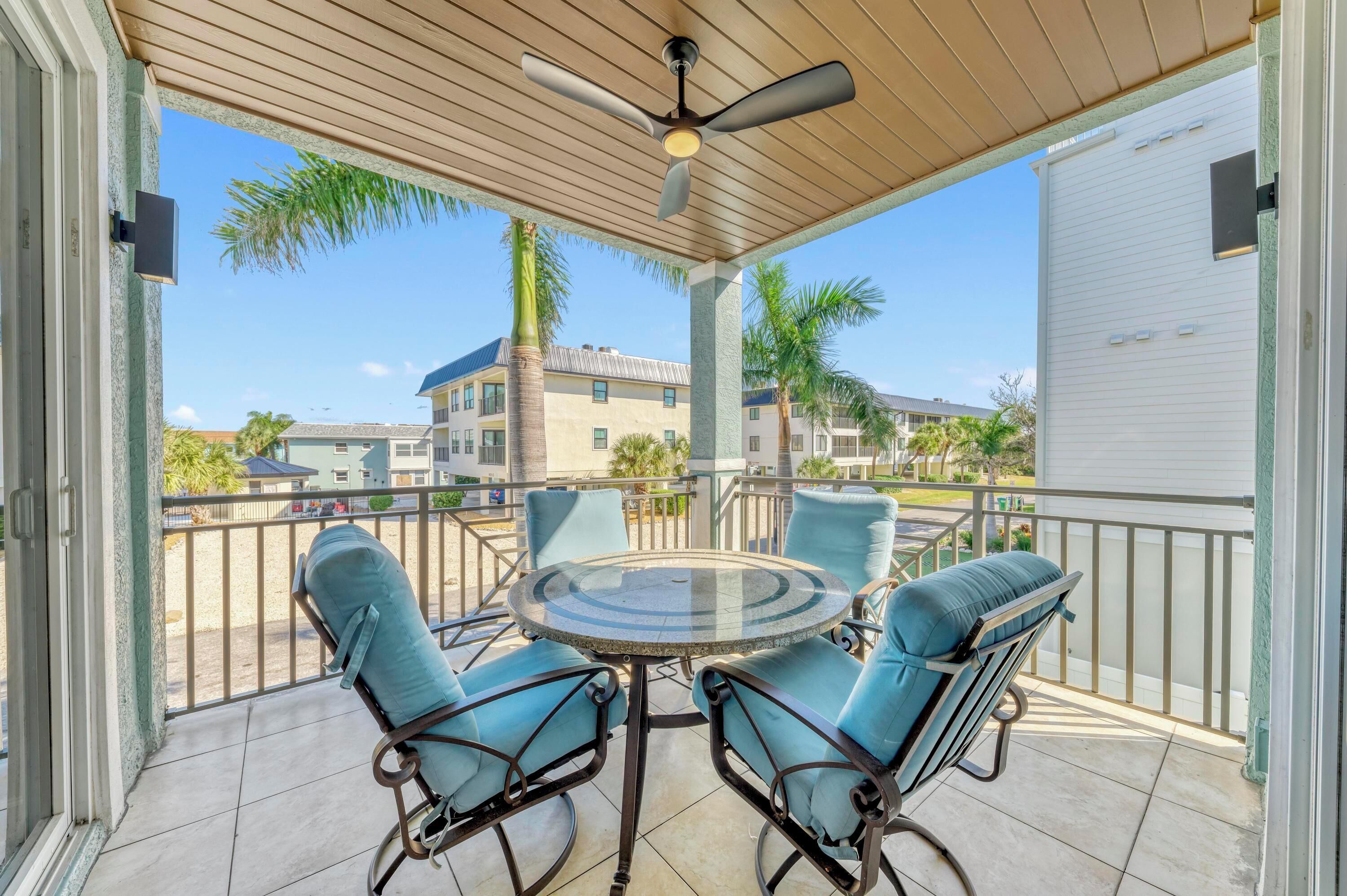 A covered patio with a round glass table and four blue cushioned chairs, a ceiling fan, and a view of neighboring houses and palm trees.
