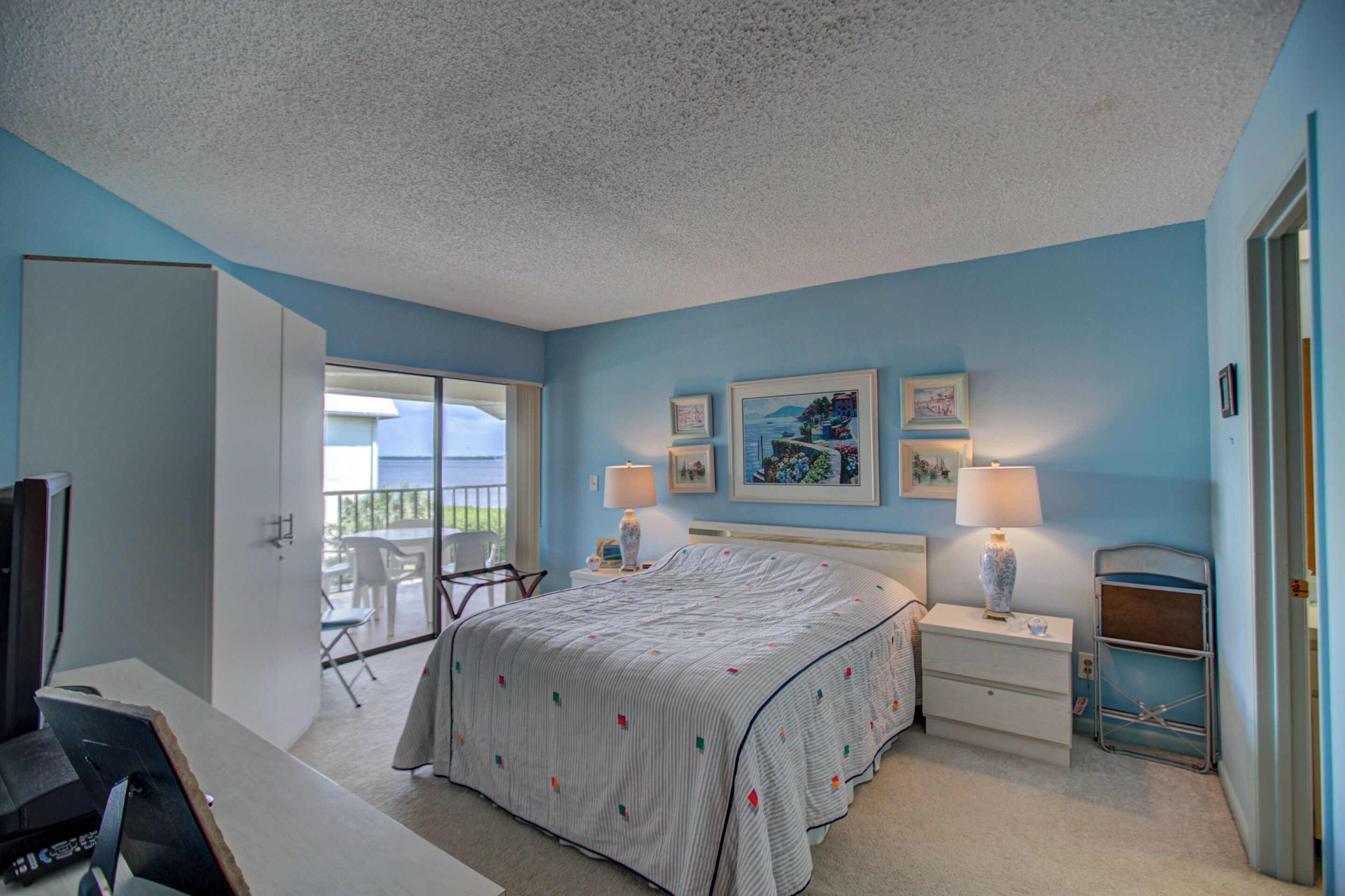 A bedroom with a bed, bedside tables, a desk, and a chair. There are two windows and a door leading to a balcony.