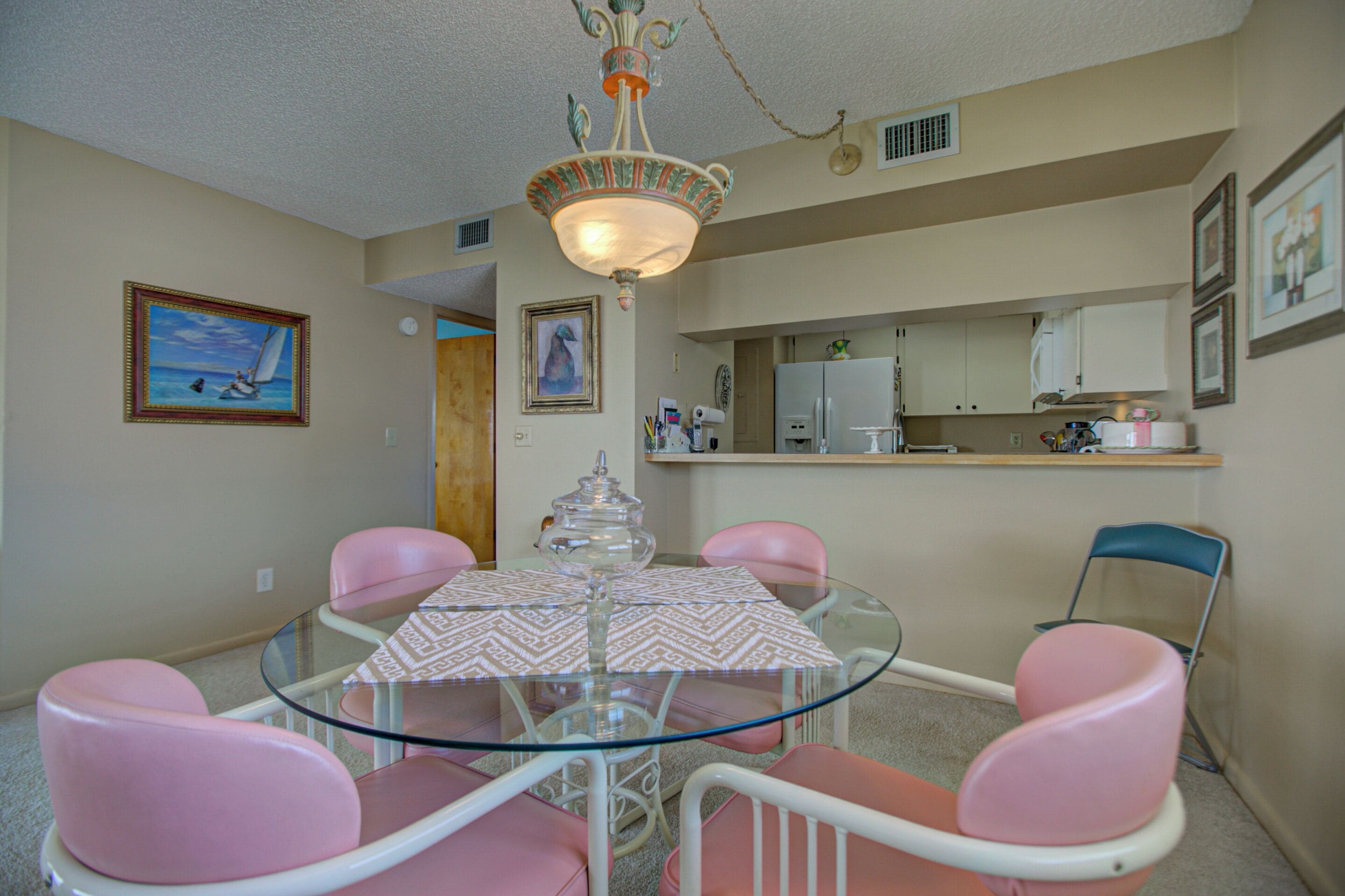 A dining area with a glass table, pink chairs, a chandelier, and a kitchen in the background.