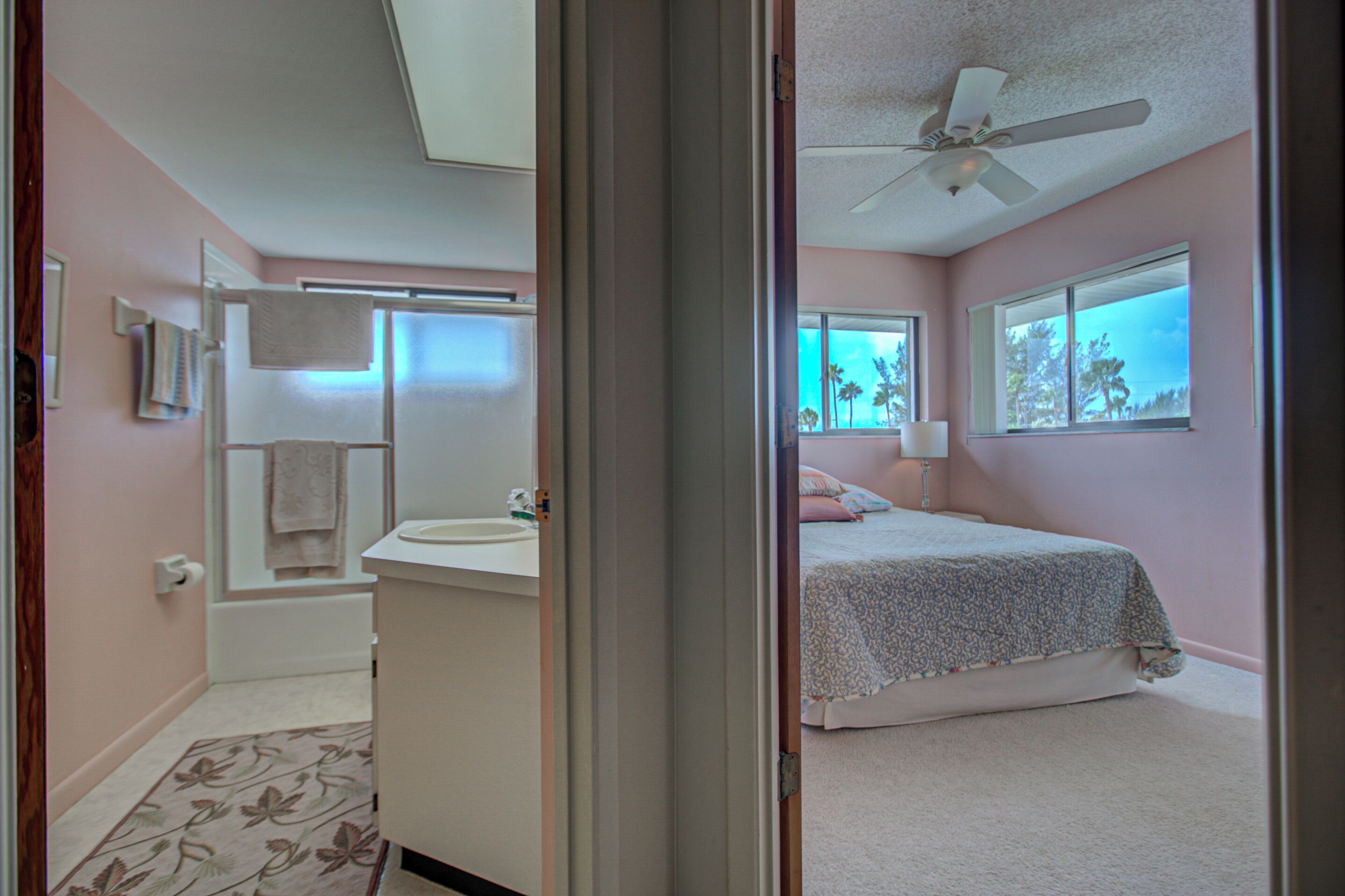 A bedroom with a bed, a window with a view, a ceiling fan, and a bathroom with a bathtub and a towel rack.