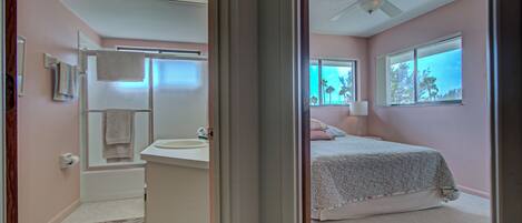 A bedroom with a bed, a window with a view, a ceiling fan, and a bathroom with a bathtub and a towel rack.