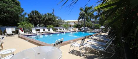 A pool area with lounge chairs and a table, surrounded by palm trees and a clear blue sky.
