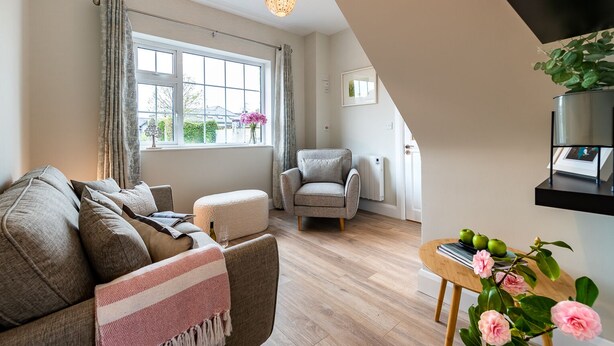A modern living room with a sofa, armchair, and ottoman, a large window with curtains, a hanging light fixture, and a small table with books and flowers. - Modern Elegant Townhouse in Historic Kenmare (Kenmare)
