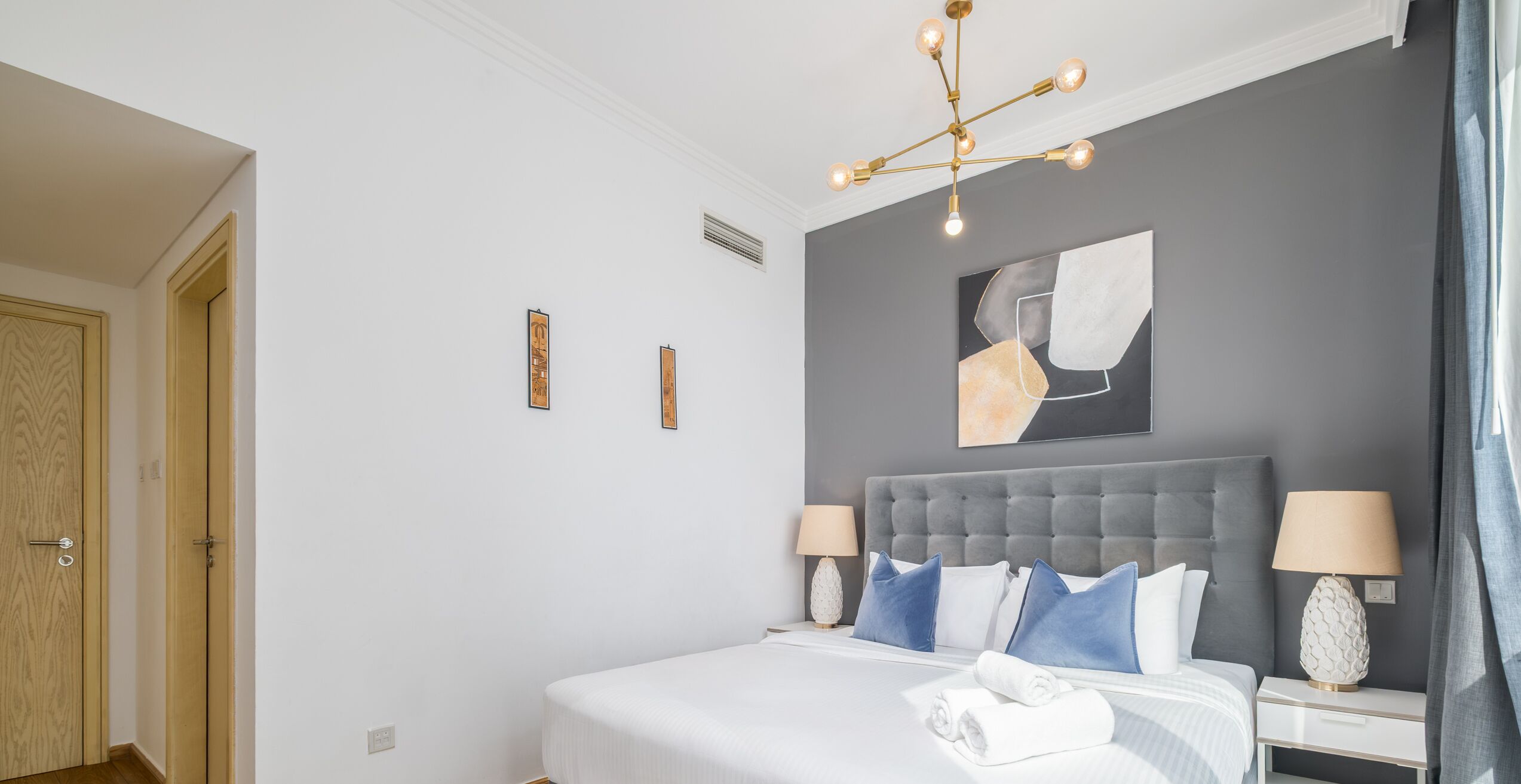 Une chambre d’hôtel moderne avec un grand lit, deux tables de chevet avec des lampes, une tête de lit rembourrée de couleur grise et un luminaire au plafond.