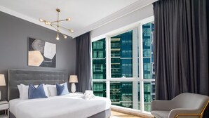 Une chambre d’hôtel moderne avec un grand lit, deux tables de chevet, un lustre, une vue sur un immeuble à travers de grandes fenêtres du sol au plafond et un fauteuil gris.