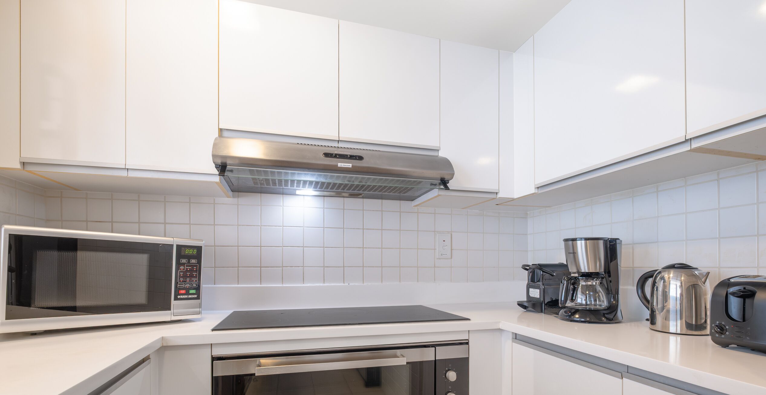 Een moderne keuken met witte kastjes, een ingebouwde magnetron, een koffiezetapparaat en een waterkoker.