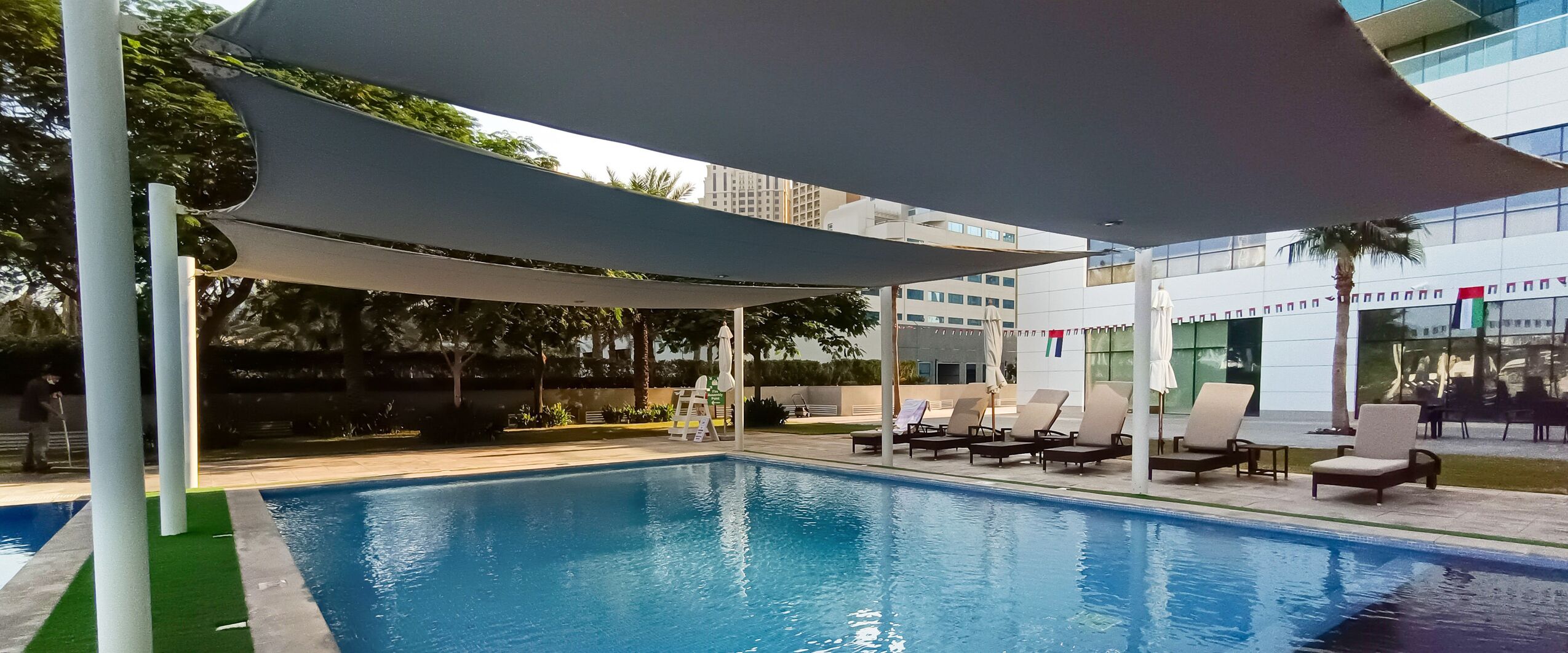 A swimming pool with lounge chairs and umbrellas, surrounded by greenery and modern buildings.