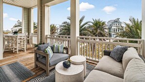 Un espace extérieur abrité doté d’une terrasse en bois, d’un canapé, d’une table à manger avec des chaises, et offrant une vue sur des palmiers et des maisons en arrière-plan.