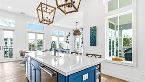 Une cuisine spacieuse dotée d’un grand îlot central, de plans de travail blancs et de meubles de rangement bleus. De nombreuses fenêtres laissent entrer la lumière naturelle et offrent une vue sur l’extérieur.