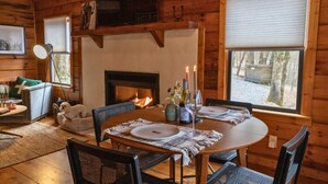 A cozy cabin interior with a fireplace, wooden walls, a dining table with chairs, and a view of the outdoors.