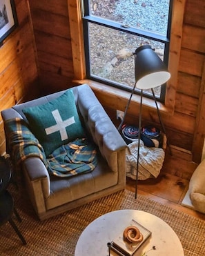 A cozy room with a wooden interior, a cushioned armchair with a cross-patterned blanket, a standing lamp, and a small round table with a book. - Designer Creekside Cabin on Fightingtown Creek | Hot Tub, SMEG Kitchen & Fishing (Cherry Log)
