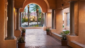 Un paseo techado con columnas, plantas en macetas y vistas a mesas al aire libre y palmeras.