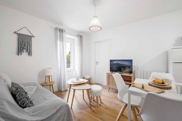 Ein modernes, minimalistisches Wohnzimmer mit einer Couch, einem Esstisch mit Stühlen, einem Fernseher und einer kleinen Küchenzeile.