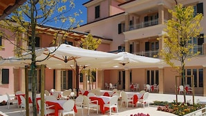 Une cour dotée d’un espace extérieur aménagé avec des chaises et des tables blanches, le tout entouré d’un bâtiment orné de balcons et doté d’une façade rose.