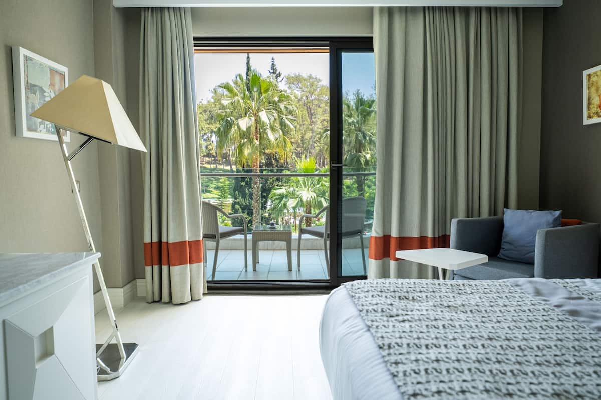 A hotel room with a bed, a desk, a chair, a table, and a balcony with a view of palm trees.