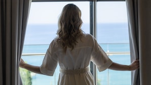Una persona con un vestido blanco de pie junto a una ventana con vista al mar.