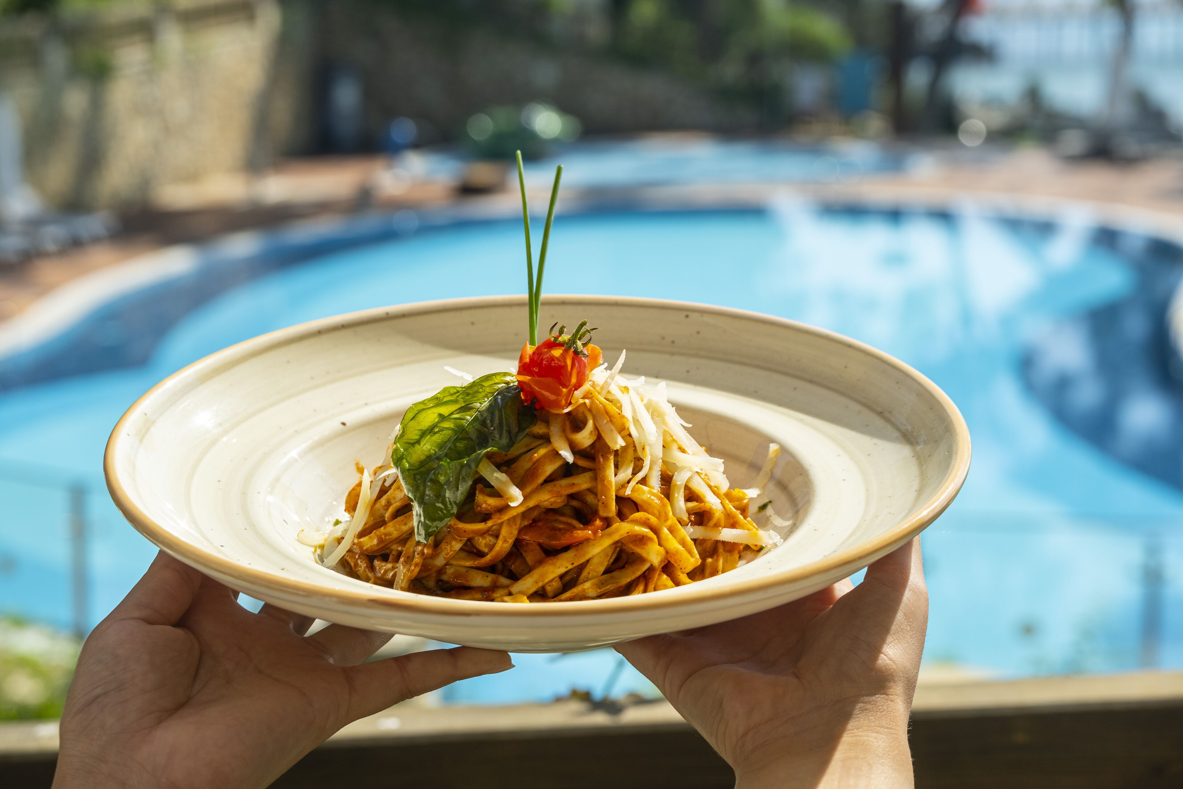 A person holding a plate of pasta with a garnish, with a pool in the background.