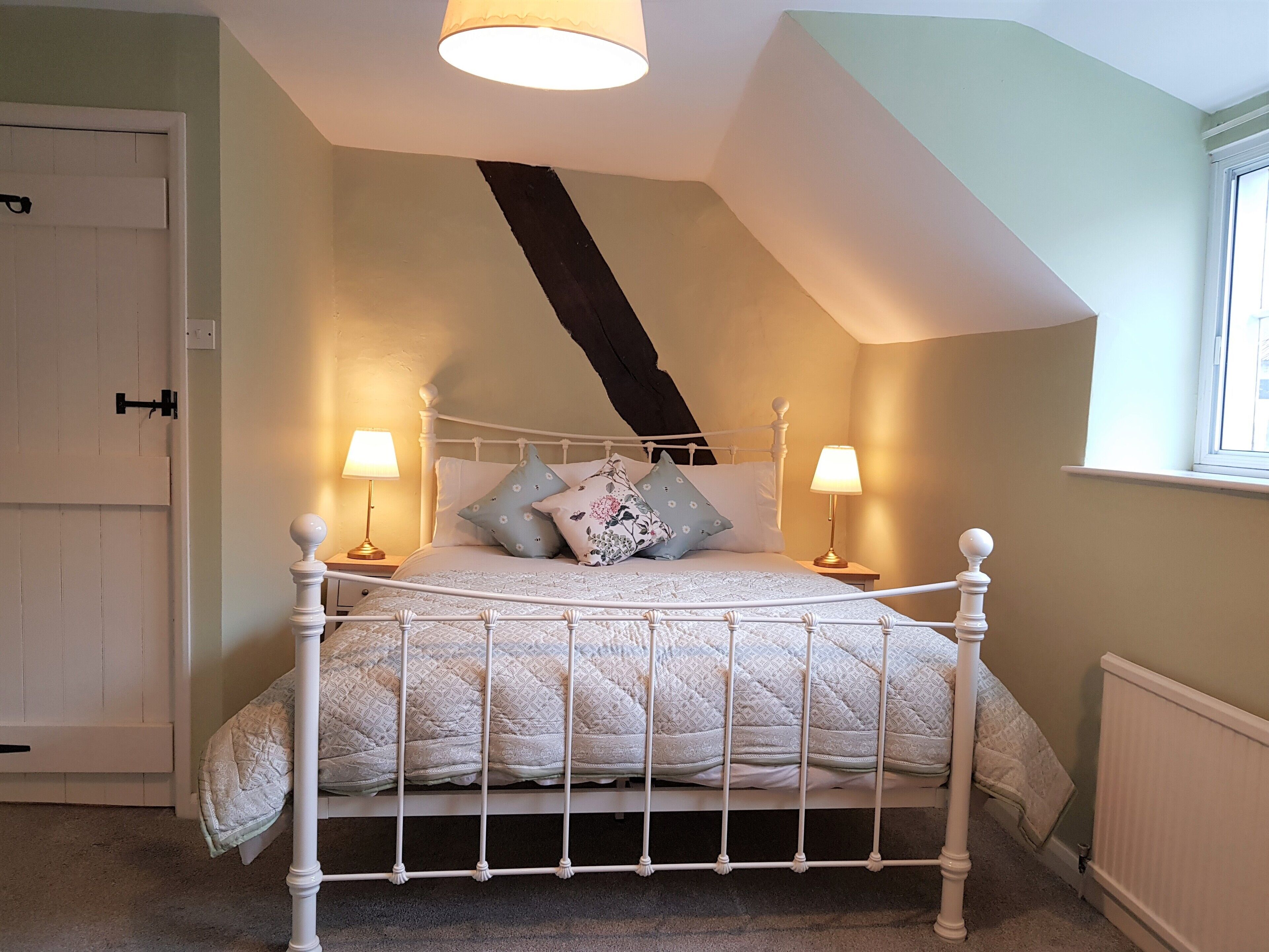 Une chambre à coucher avec un lit en métal blanc, deux lampes de chevet, une fenêtre donnant sur un beau paysage et un plafond en pente.