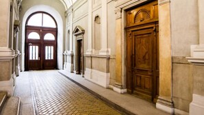 Un couloir doté d’un sol carrelé, d’une porte en bois surmontée d’un tympan et, au bout, d’une grande porte en arc de cercle.