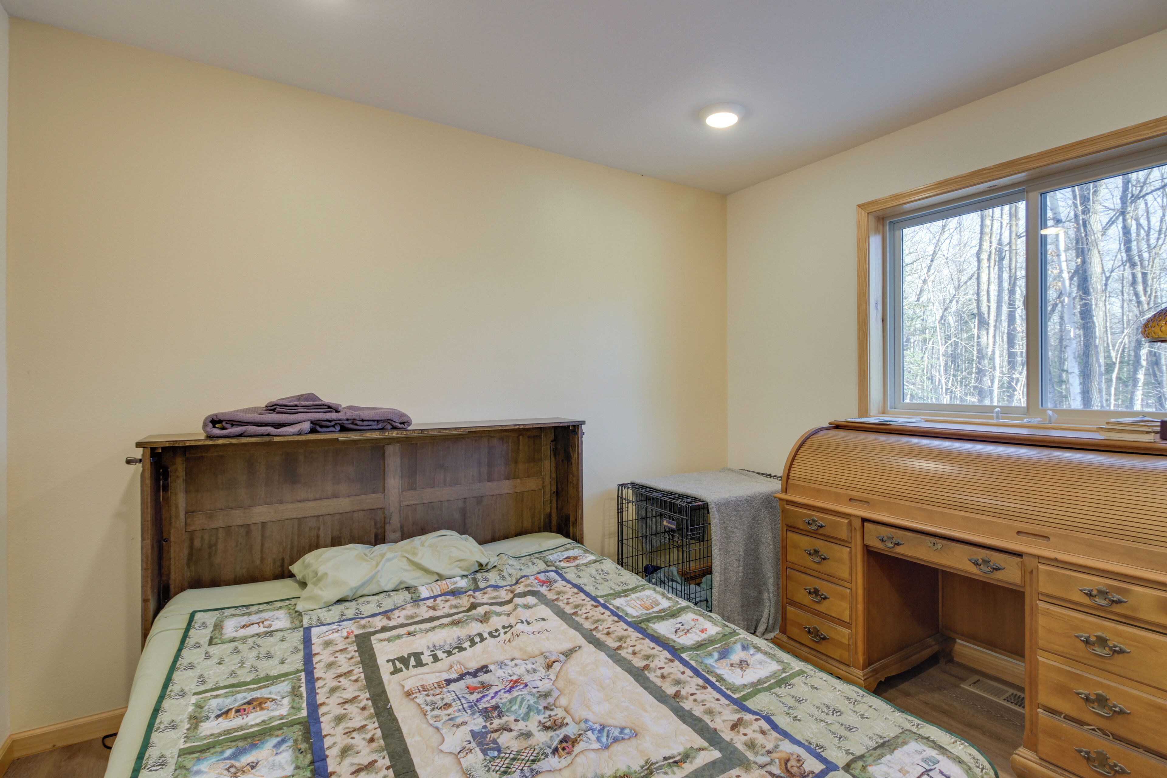 Une chambre à coucher avec un lit, un bureau en bois, une fenêtre donnant sur des arbres et une courtepointe ornée d’un motif de carte géographique.