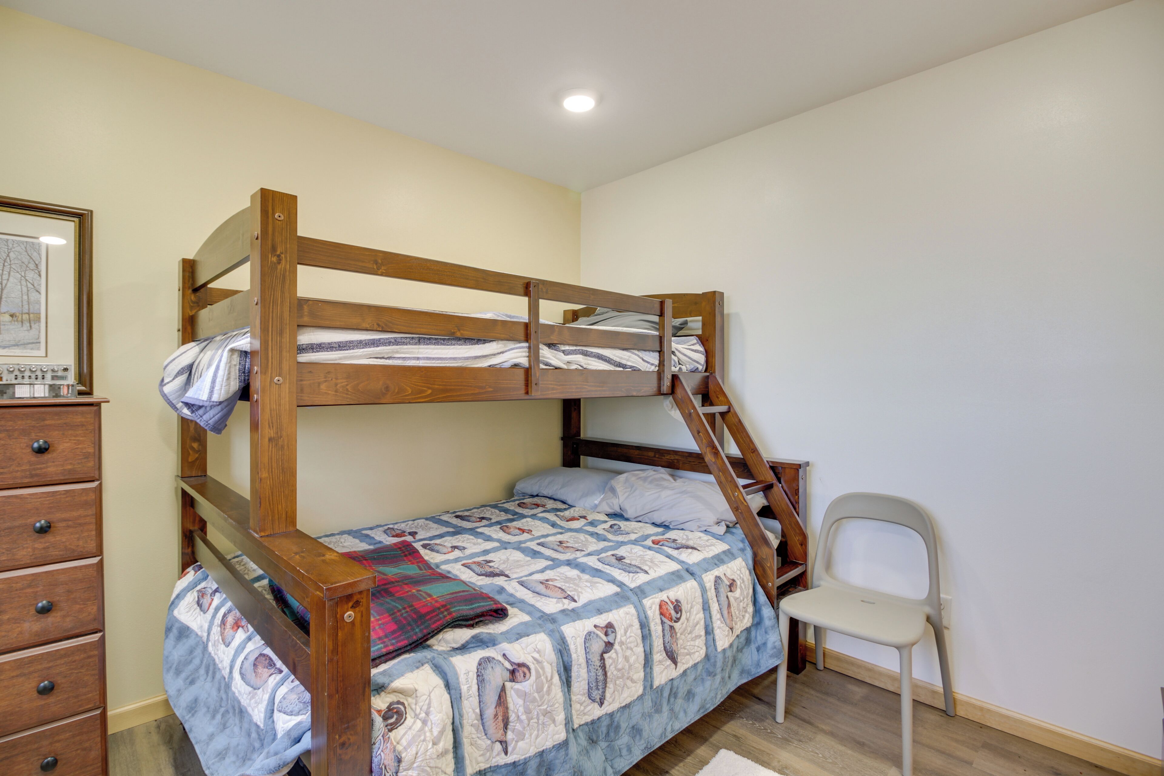 A bunk bed with a wooden frame, a dresser with drawers, a chair, and a framed picture on the wall.
