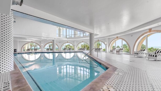 A modern indoor swimming pool with clear blue water, surrounded by white tiled floors and walls, and furnished with white lounge chairs and umbrellas.