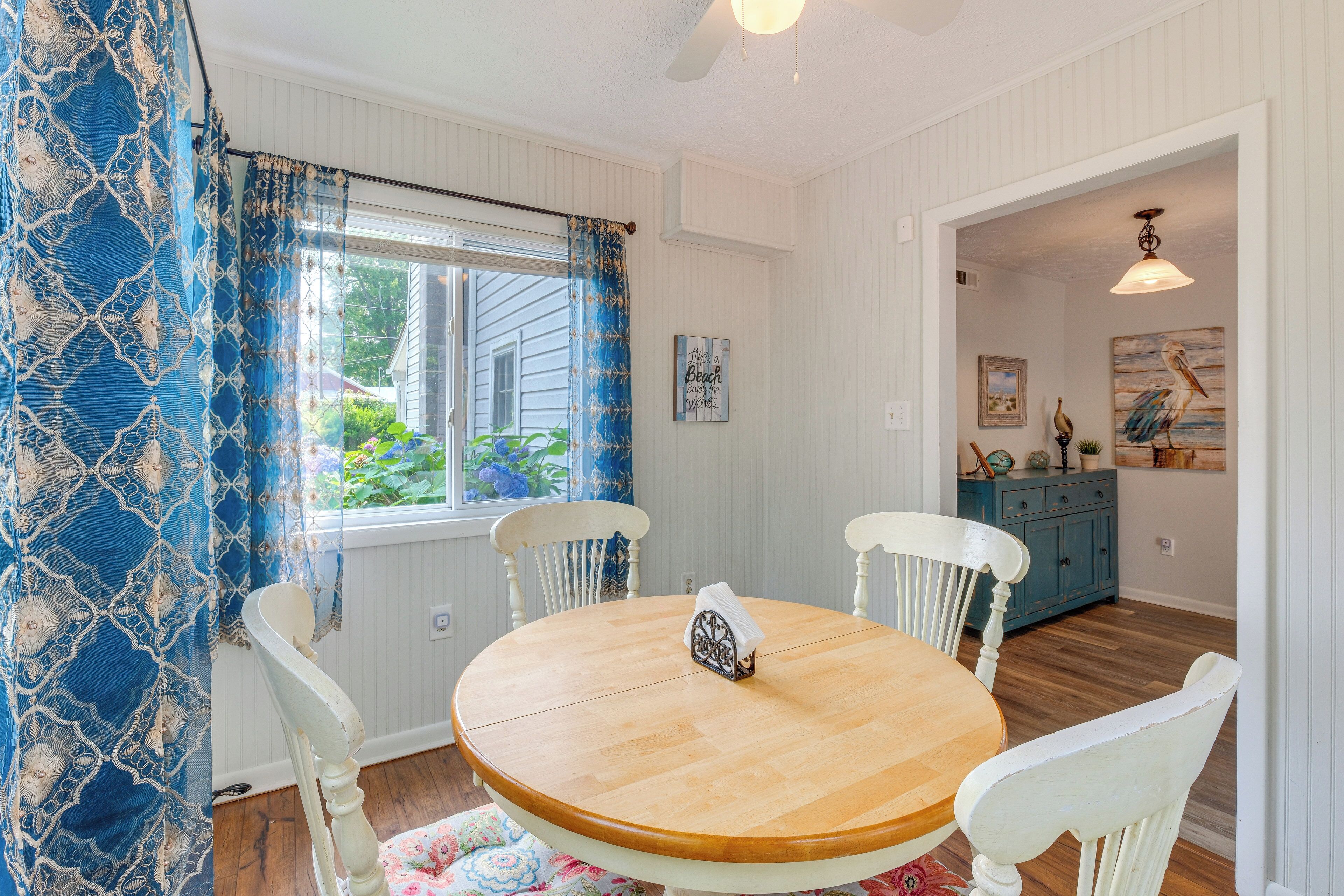 A dining area with a round wooden table, white chairs, and a window with blue curtains.