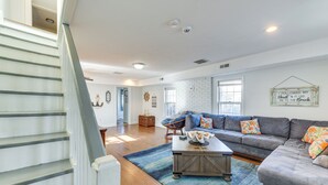 A spacious living room with a gray sectional sofa, a coffee table, and a blue rug. A staircase with white railings leads to another room. - Adorable Cottage: Fire Pit, Walk to Colonial Beach (Colonial Beach)