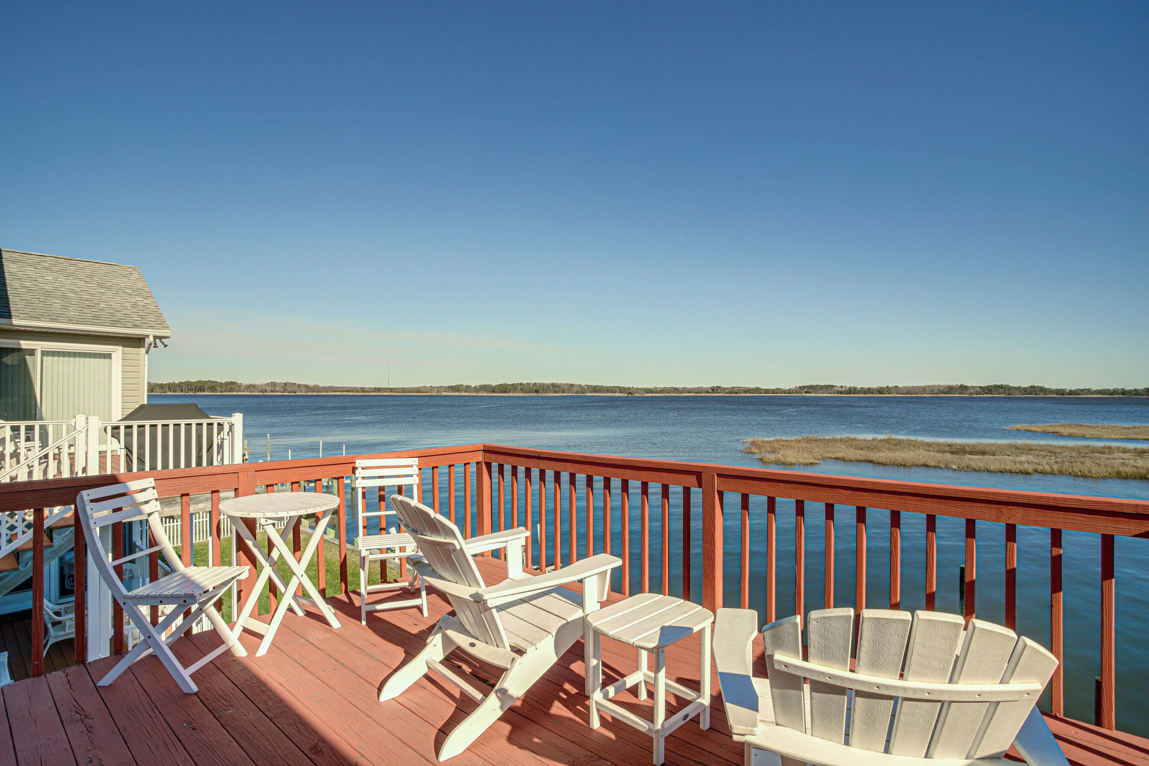A wooden deck with white Adirondack chairs and a table overlooking a body of water.