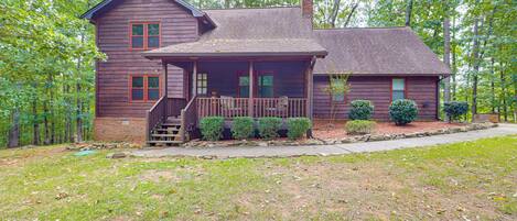 A two-story wooden house with a porch, surrounded by trees and a grassy yard.