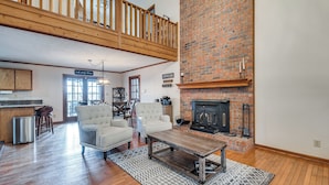 A spacious living room with a high ceiling, a fireplace, and a kitchen area visible in the background.