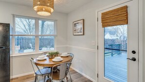 A dining area with a wooden table, metal chairs, a hanging light fixture, and a view through a window with blinds.