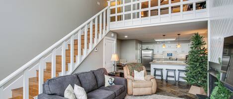 Un espacio habitable amplio con un sofá gris, un sillón color beige, un pequeño árbol de Navidad y una escalera que lleva a un nivel superior.
