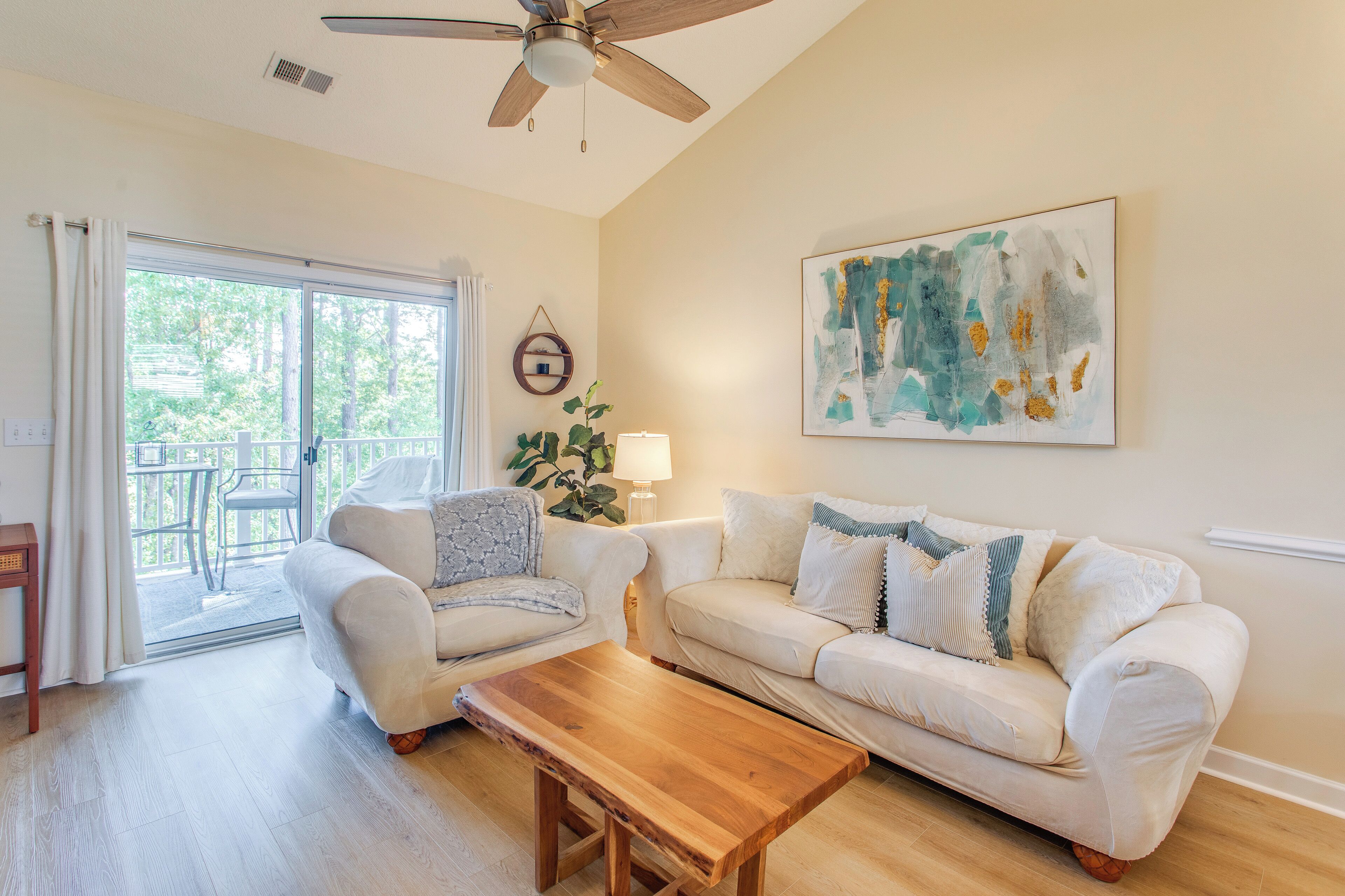 A living room with a sofa, armchair, and coffee table, a ceiling fan, a painting, and a view through a glass door.