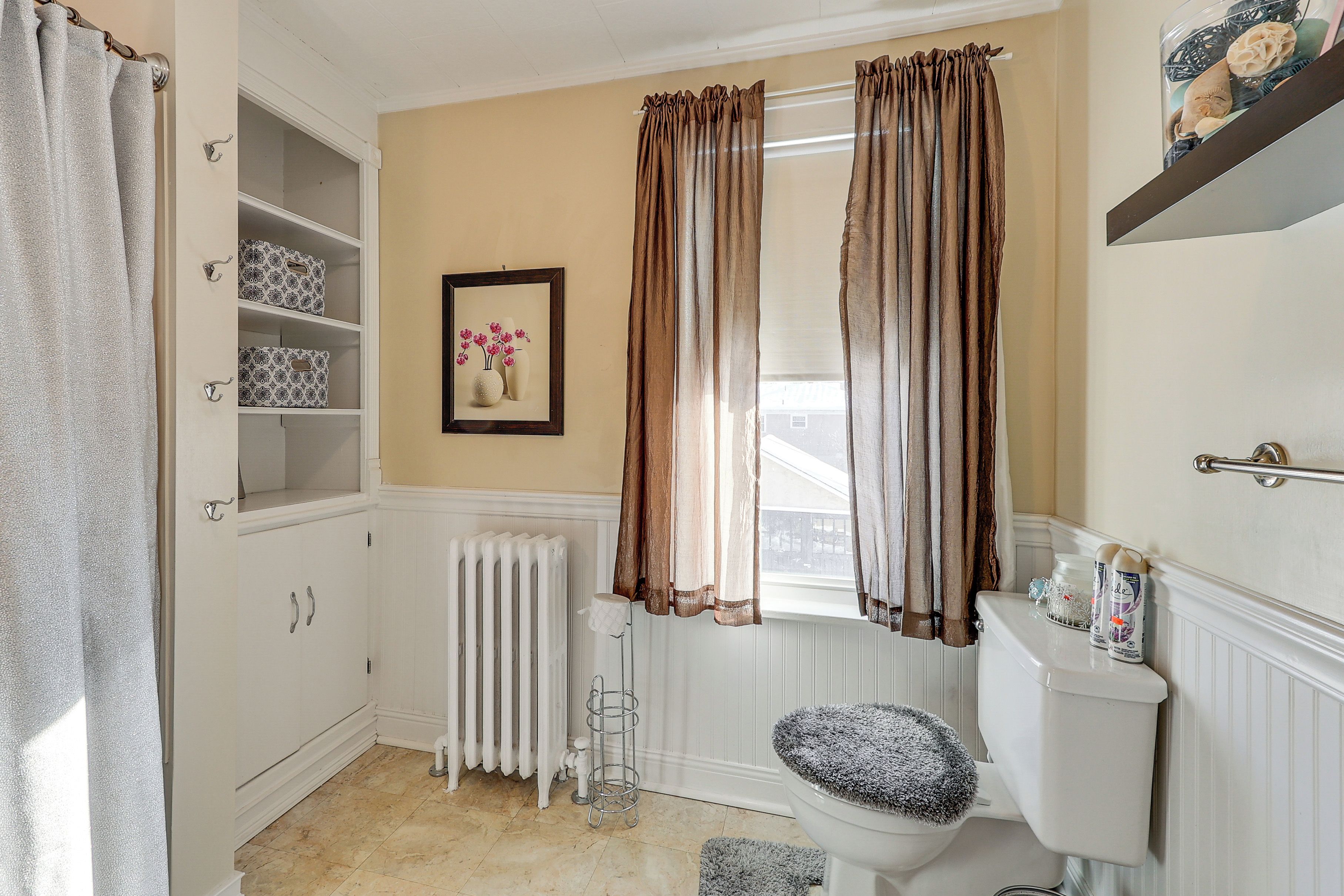 A bathroom with a toilet, radiator, storage cabinets, and a window with curtains.