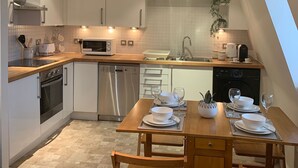A modern kitchen with white cabinets, a wooden dining table set for two, and a dishwasher. - Riding Gate Apartment (Canterbury)