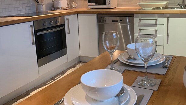 A modern kitchen with white cabinetry, a microwave, an oven, and a sink. There is a dining table set with plates, bowls, and wine glasses. - Riding Gate Apartment (Canterbury)