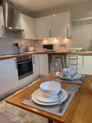 Une cuisine moderne avec des meubles blancs, un four à micro-ondes, un four et un évier. On y trouve une table à manger mise avec des assiettes, des bols et des verres à vin.