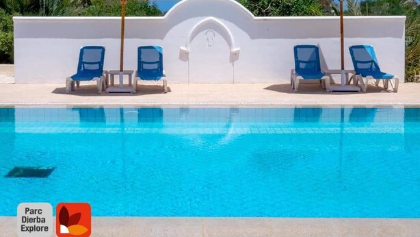 A swimming pool with blue water, surrounded by white walls and blue lounge chairs. There are two umbrellas providing shade. - Parc Djerba Explore Residence (Aghir)