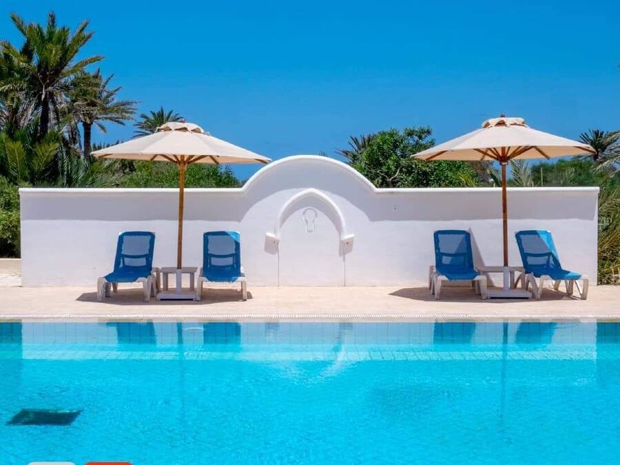 A swimming pool with blue water, surrounded by white walls and blue lounge chairs. There are two umbrellas providing shade.