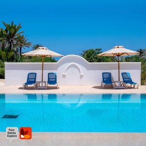 A swimming pool with blue water, surrounded by white walls and blue lounge chairs. There are two umbrellas providing shade. - Parc Djerba Explore Residence (Aghir)
