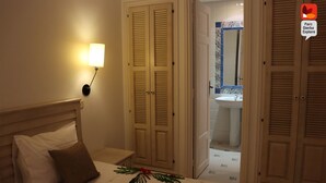 A bedroom with a bed, pillows, a nightstand, a wall-mounted lamp, and a door leading to a bathroom with a sink and mirror. - Parc Djerba Explore Residence (Aghir)
